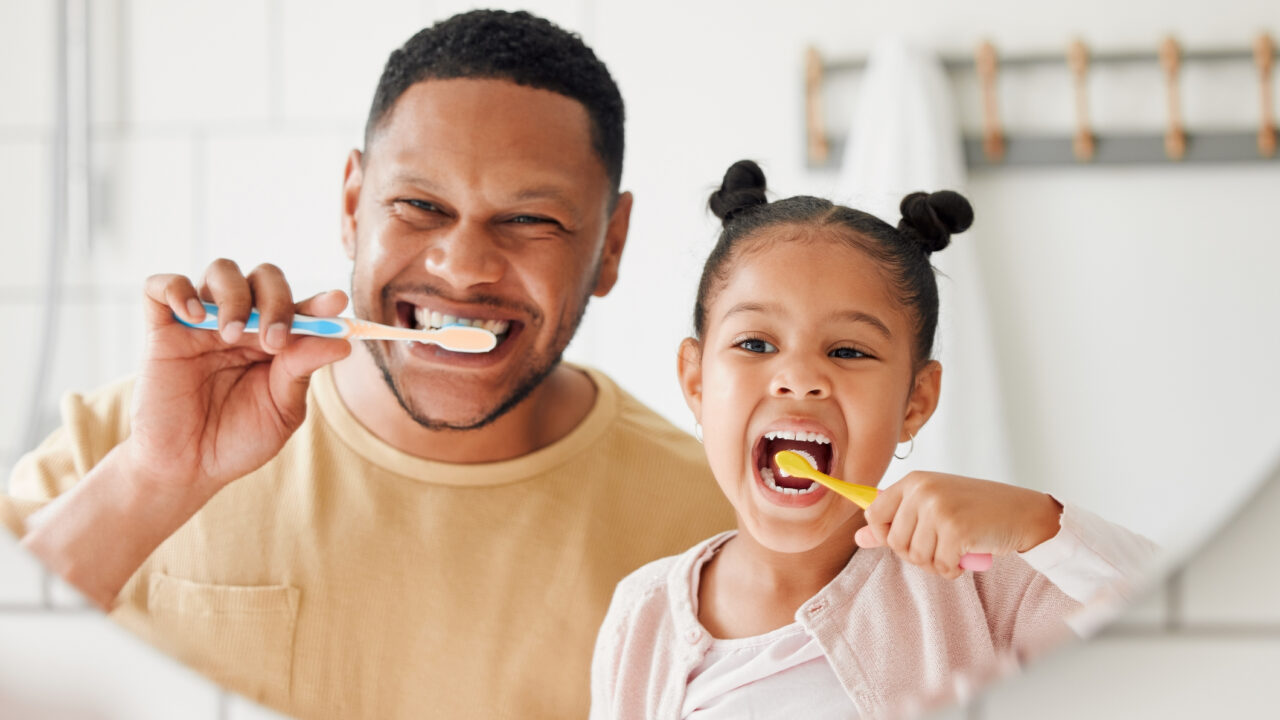 Happy mixed race father and daughter brushing their teeth together in a bathroom at home. Single African American parent teaching his daughter to protect her teeth